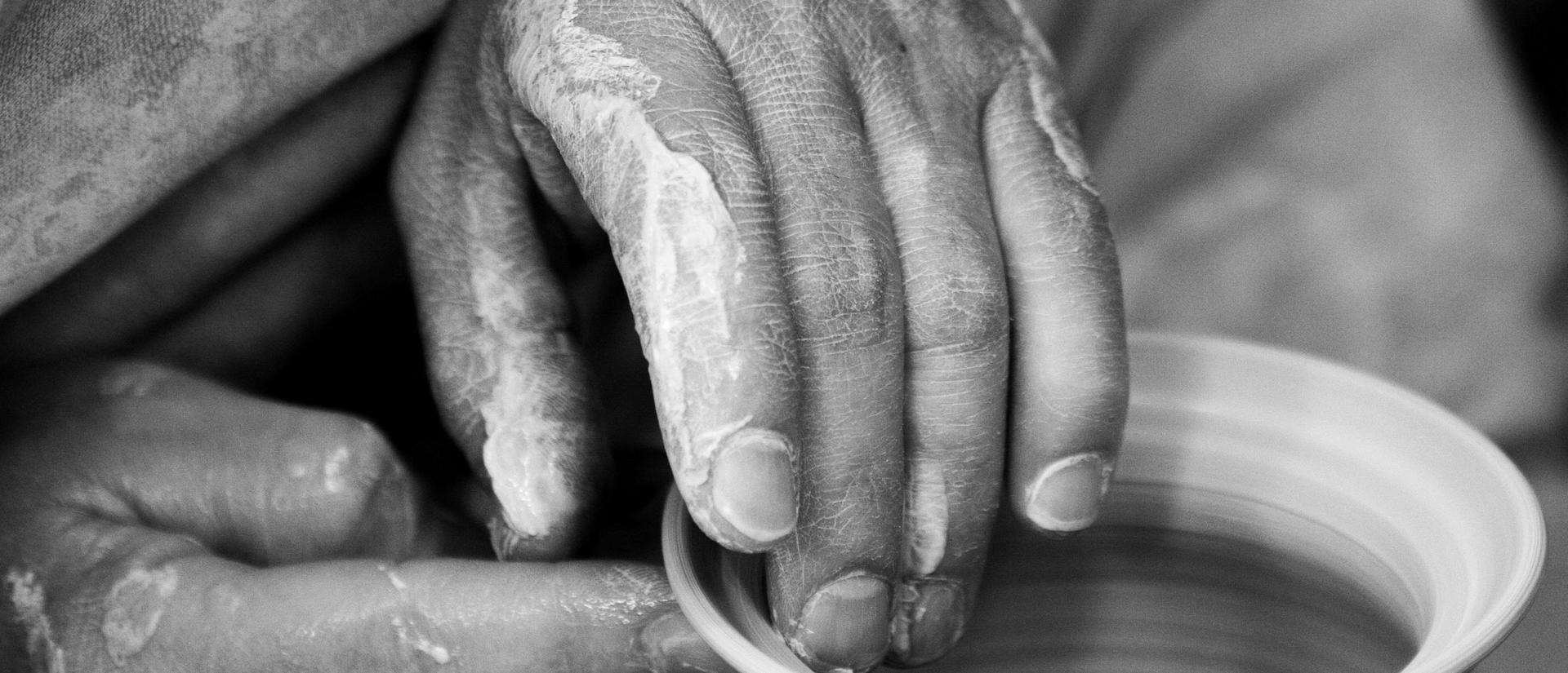 A black and white photograph of two hands throwing a ceramic pot on a wheel